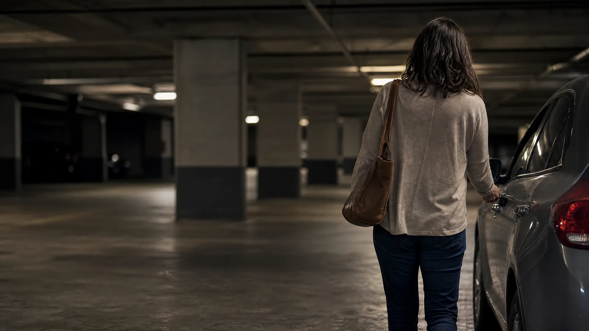 A woman seen from behind with a shoulder bag and keys stands beside a car in a dimly lit underground parking garage with concrete columns.